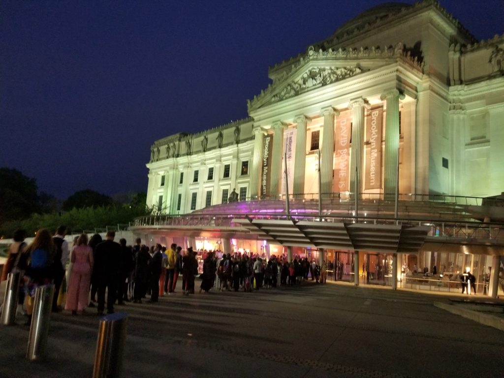 Evening picture of the front of the Brooklyn Museum with a line of people waiting to enter