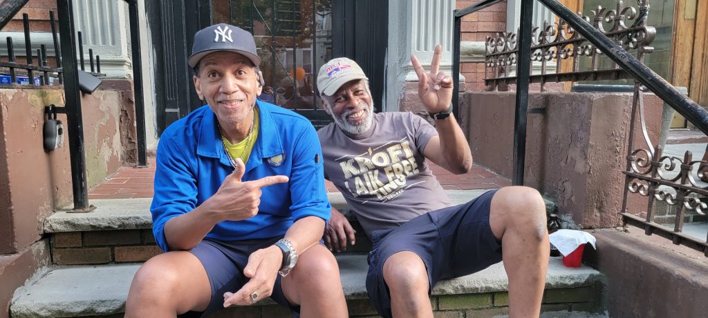 Two male neighbors sitting on wearing caps and shorts. One neighbor is giving the peace sign with his fingers.