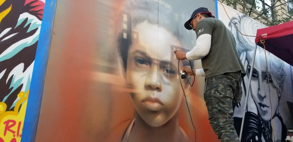Man wearing camp pants and t shirt spray painting an image of a young boy as part of a street mural